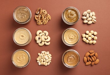 Many tasty nut butters in jars and nuts on brown table, flat lay