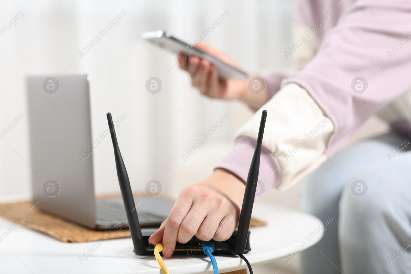 Photo of Woman with smartphone connecting cable to Wi-Fi router at table indoors, closeup