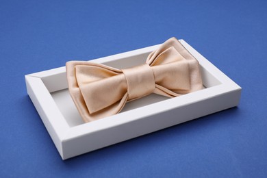 Stylish beige bow tie in box on blue background, closeup