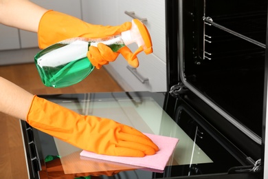 Photo of Woman cleaning electric oven with rag and detergent in kitchen, closeup