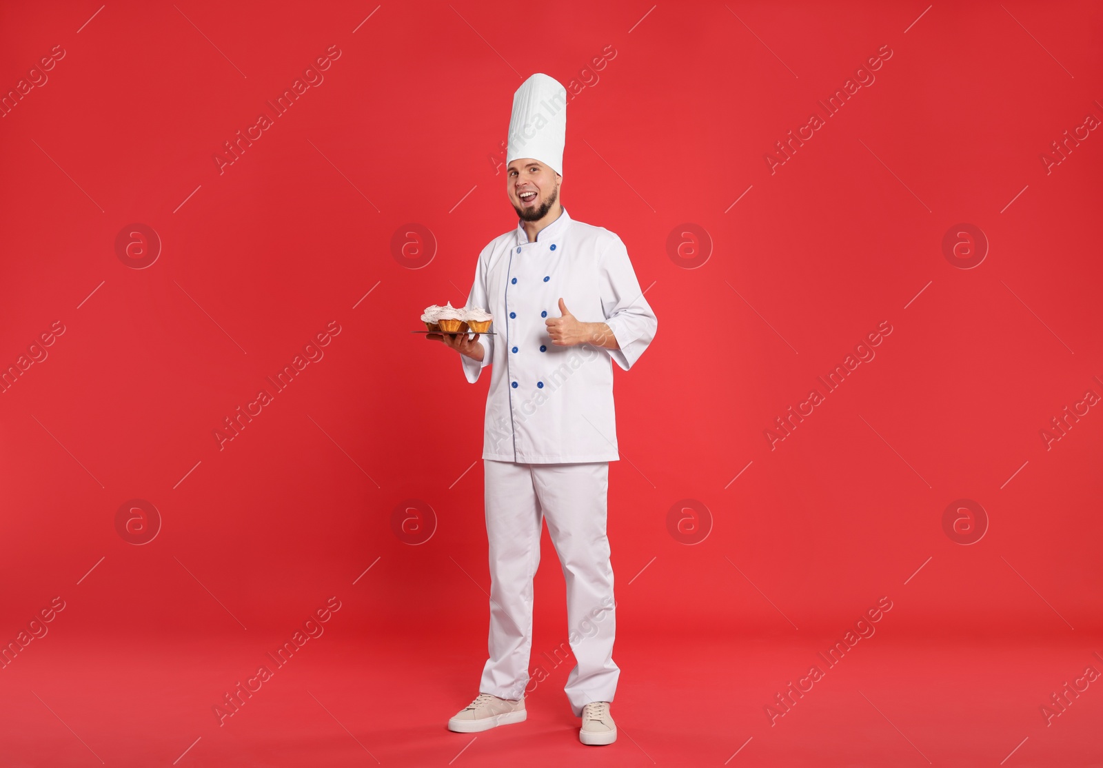 Photo of Happy professional confectioner in uniform holding delicious cupcakes and showing thumbs up on red background