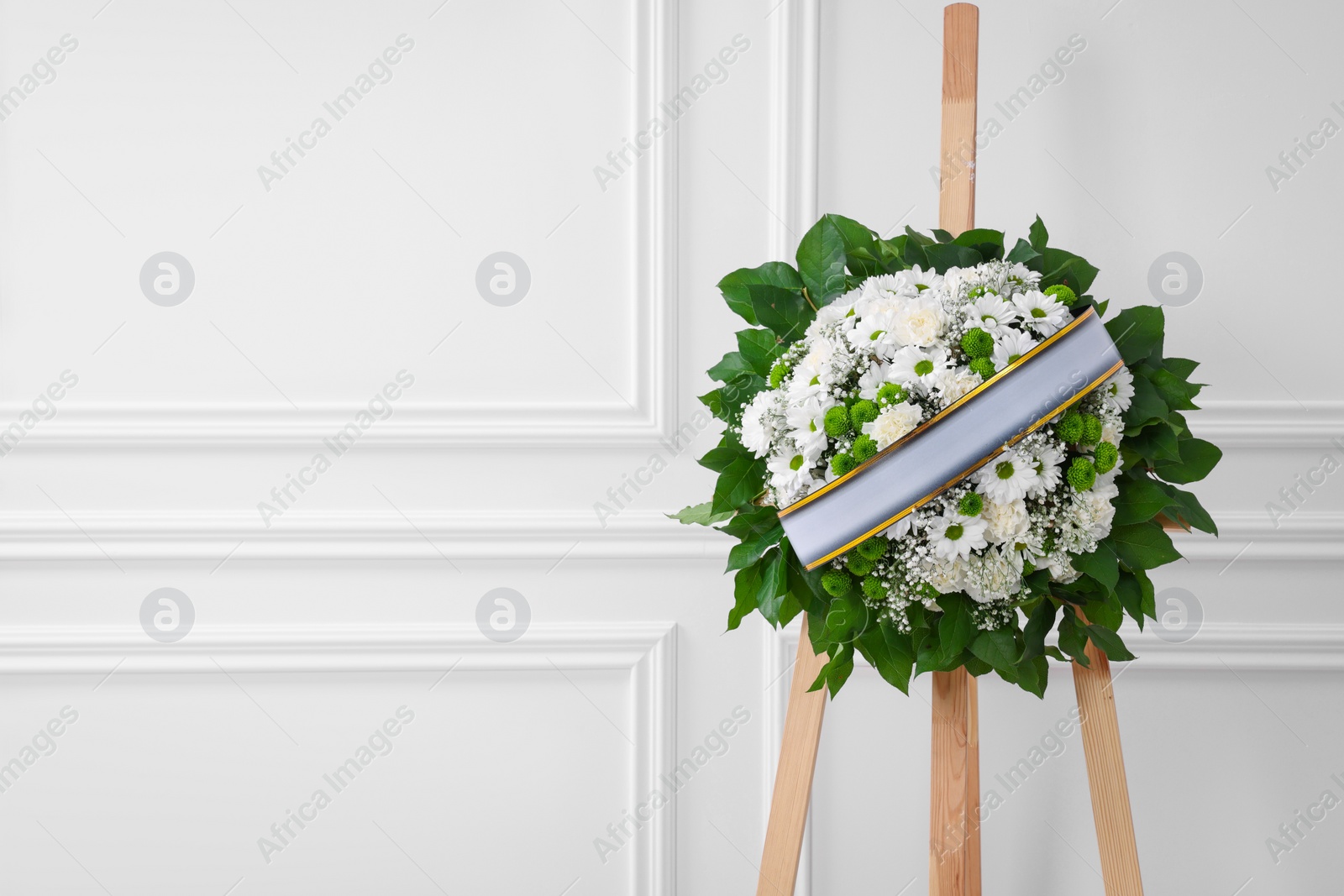 Photo of Funeral wreath of flowers with ribbon on wooden stand near white wall indoors. Space for text