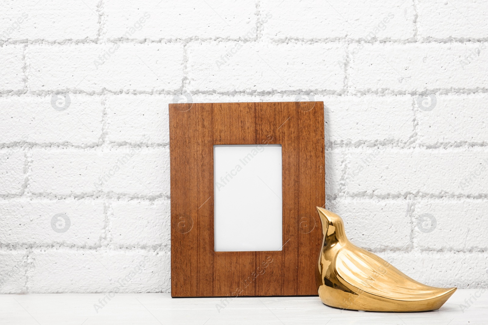 Photo of Blank frame and decorative bird figure on table near brick wall. Mock up for design