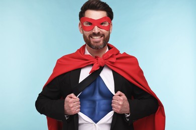 Happy businessman wearing superhero costume under suit on light blue background