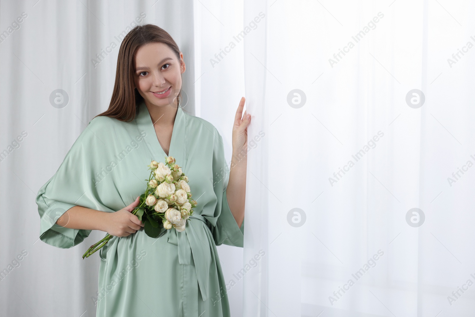 Photo of Beautiful pregnant woman with bouquet of roses near window indoors, space for text