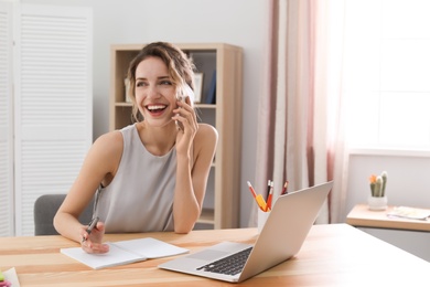 Photo of Young woman talking on mobile phone while working with laptop at desk. Home office