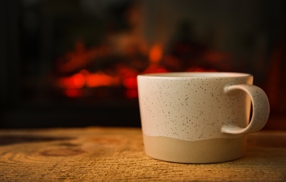 Cup with hot drink on wooden table against fireplace, space for text