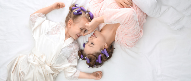 Image of Happy mother and daughter with curlers lying on bed, top view. Banner design