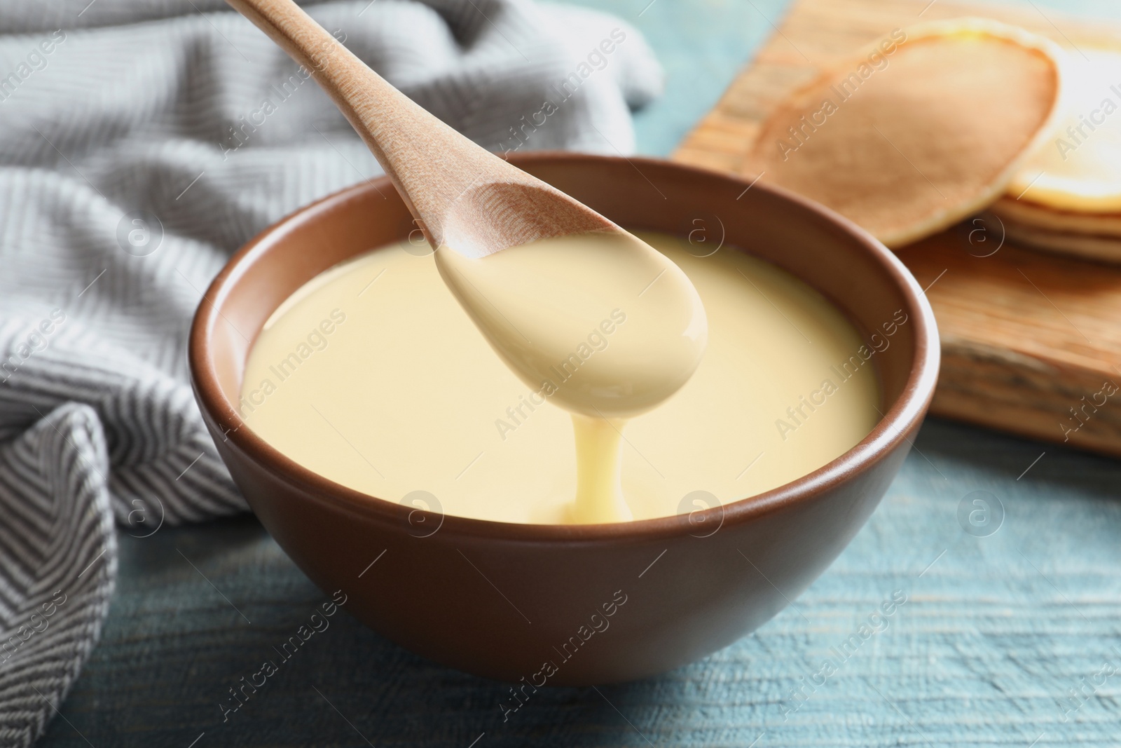 Photo of Spoon of pouring condensed milk over bowl on table, closeup. Dairy products