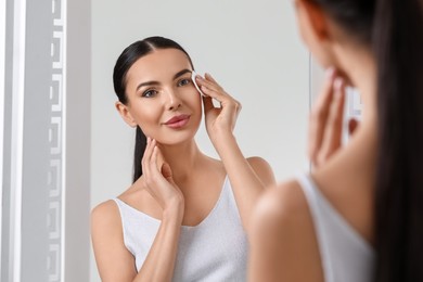 Photo of Beautiful woman removing makeup with cotton pad near mirror indoors