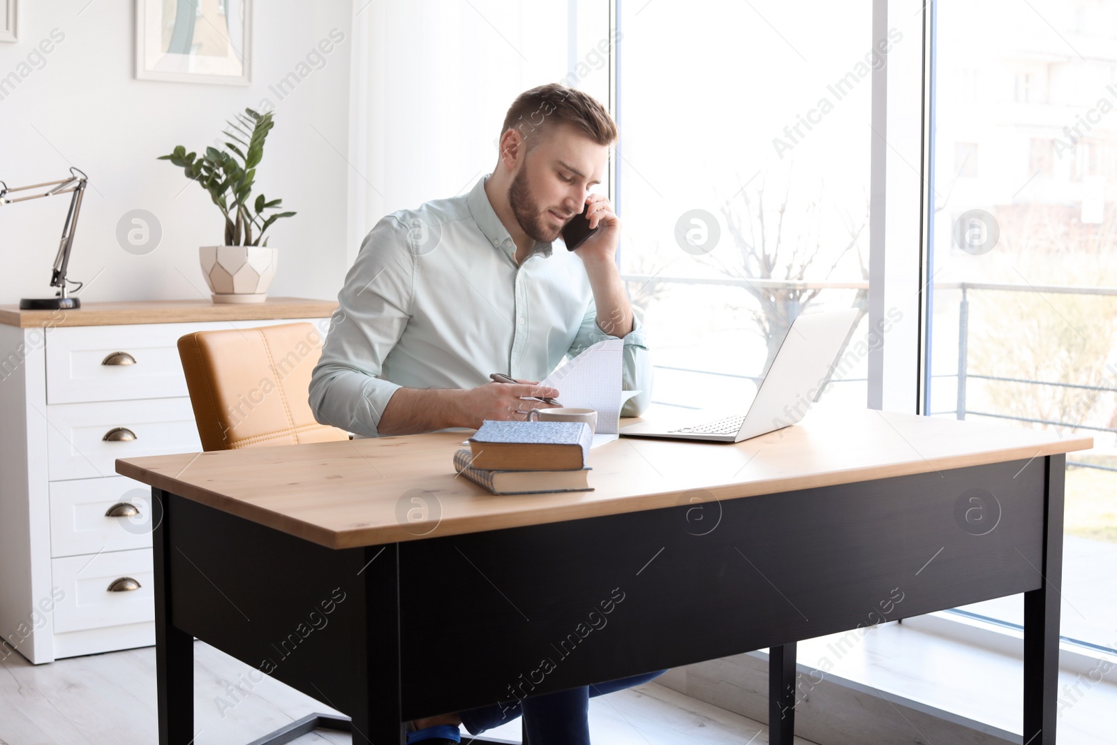 Photo of Young man talking on mobile phone while working with laptop at desk. Home office