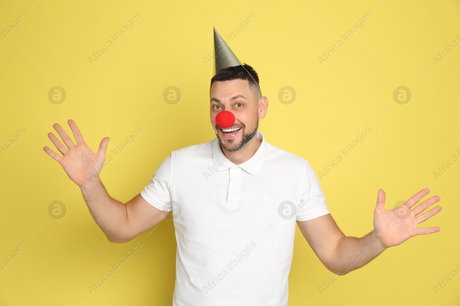 Photo of Funny man with clown nose and party hat on yellow background. April fool's day