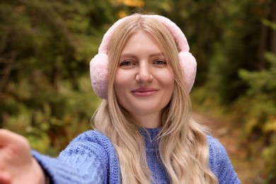 Photo of Young woman in warm earmuffs taking selfie outdoors on autumn day