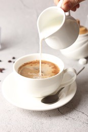 Photo of Woman pouring milk into cup with coffee at light grey textured table, closeup