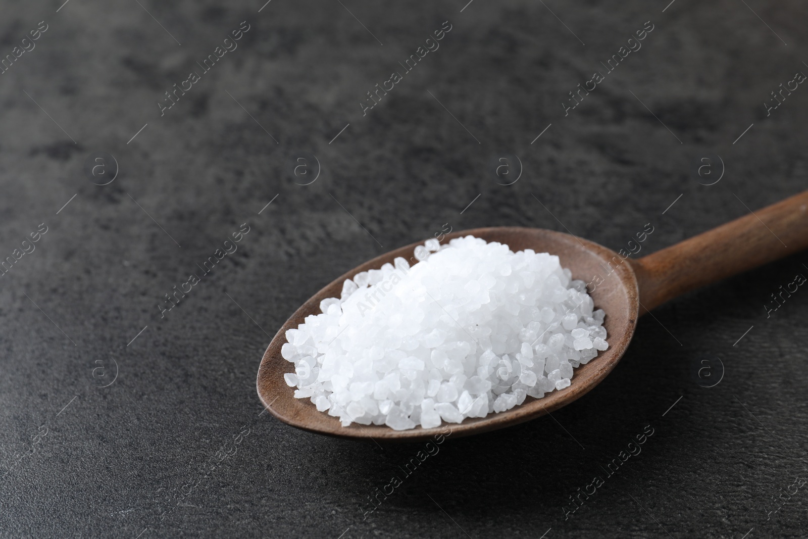 Photo of Natural salt in wooden spoon on dark grey table, closeup. Space for text