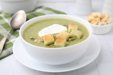 Photo of Delicious broccoli cream soup with croutons, sour cream and pumpkin seeds served on white tiled table, closeup