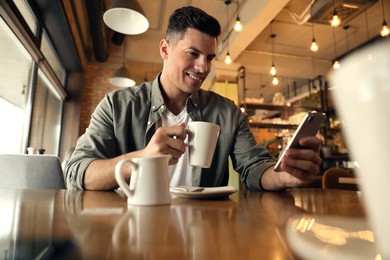 Photo of Handsome man with cup of coffee and smartphone at cafe in morning