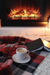 Cup of hot drink and book on blanket near fireplace indoors. Magic atmosphere