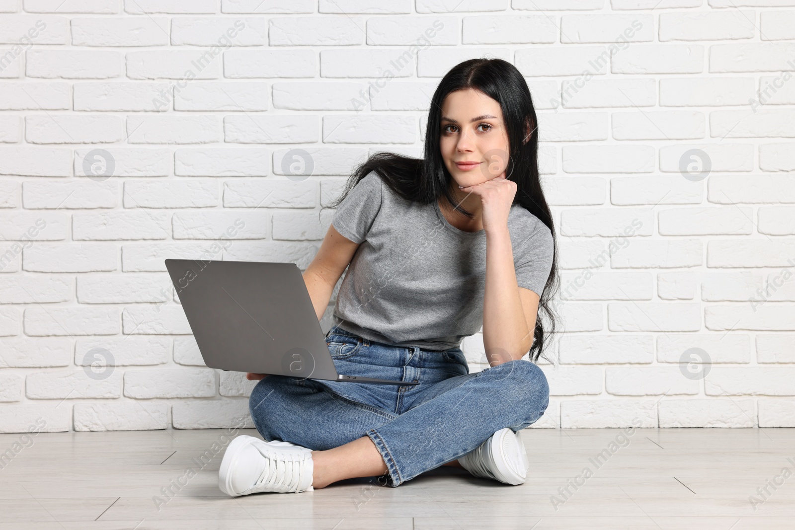 Photo of Student with laptop sitting on floor near white brick wall