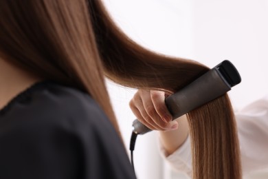 Photo of Hairdresser straightening woman's hair with flat iron indoors, closeup