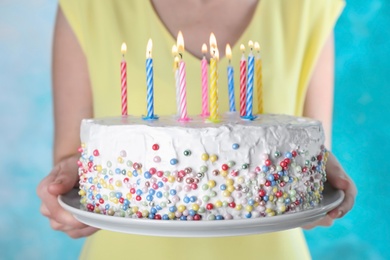 Woman holding birthday cake with burning candles on light blue background, closeup