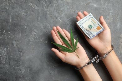 Woman in handcuffs holding hemp leaf and dollars on grey stone background, closeup. Space for text