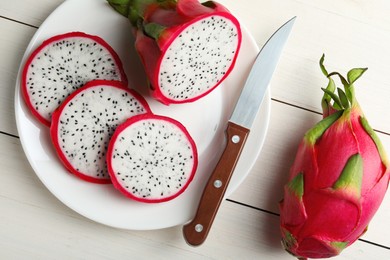 Photo of Plate of delicious cut and whole pitahaya fruits with knife on white wooden table, flat lay