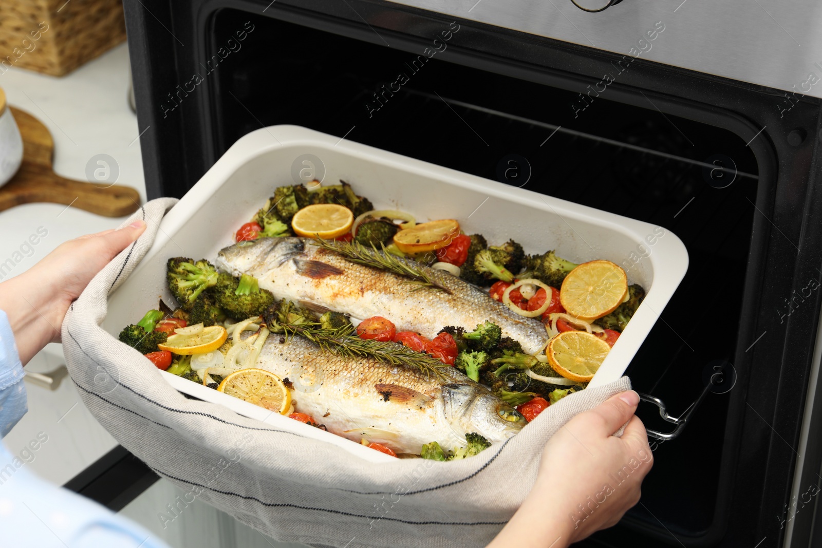 Photo of Woman taking baking dish with delicious fish and vegetables from oven in kitchen, closeup