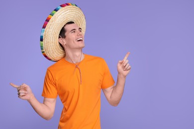 Young man in Mexican sombrero hat pointing at something on violet background. Space for text