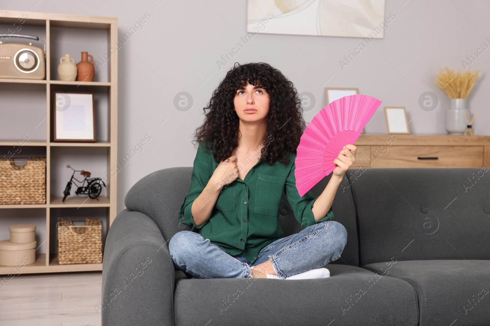 Photo of Young woman waving pink hand fan to cool herself on sofa at home