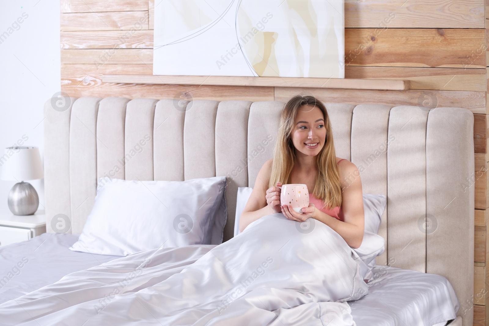 Photo of Young woman holding cup of drink on comfortable bed with silky linens, space for text