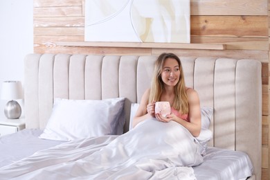 Young woman holding cup of drink on comfortable bed with silky linens, space for text