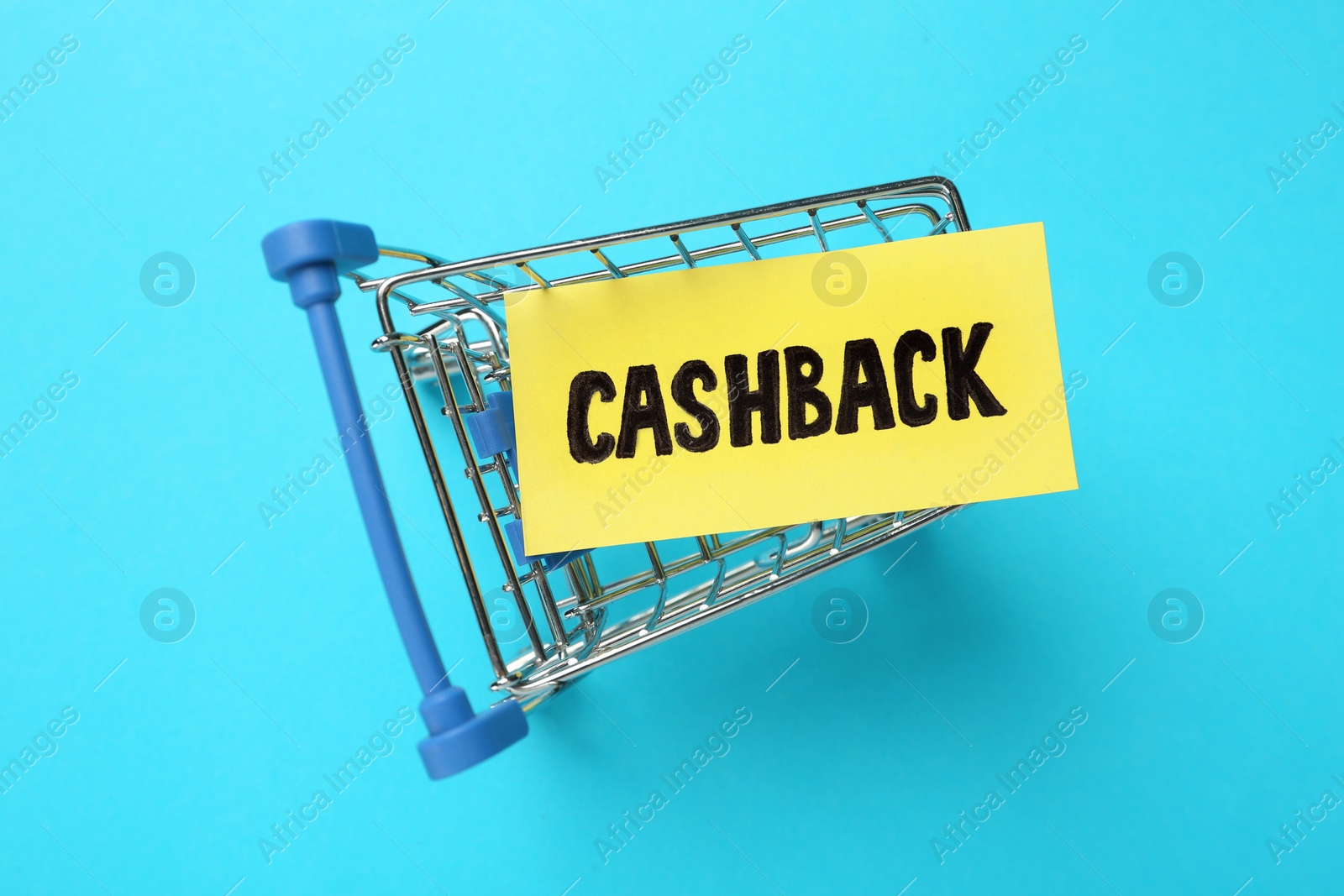 Photo of Card with word Cashback in shopping cart on light blue background, top view