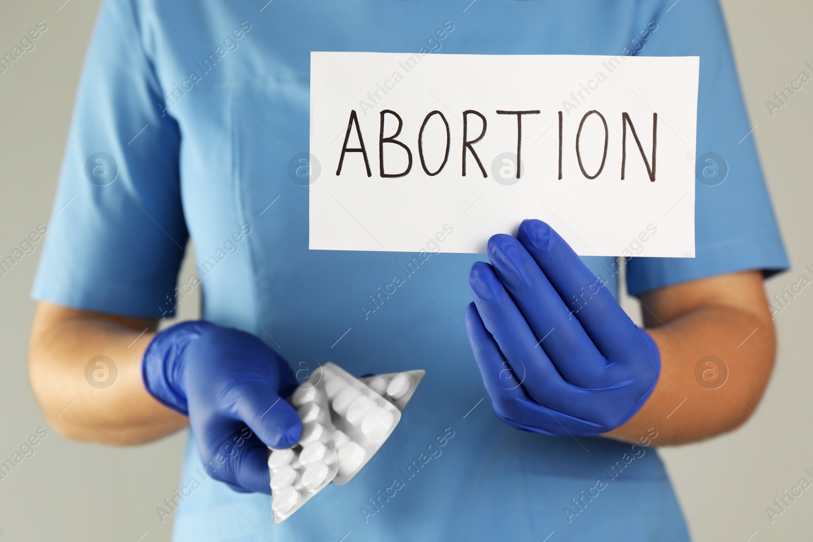 Photo of Doctor holding pills and paper card with word ABORTION on light grey background, closeup