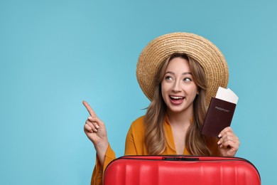 Photo of Happy young woman with passport, ticket and suitcase pointing at something on light blue background, space for text