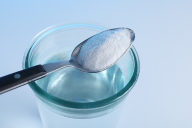 Photo of Glass of water and spoon with baking soda on light blue background, closeup