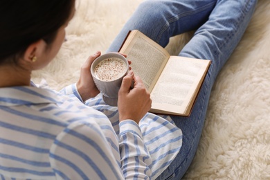 Photo of Woman with cup of coffee reading book on fuzzy rug at home, closeup