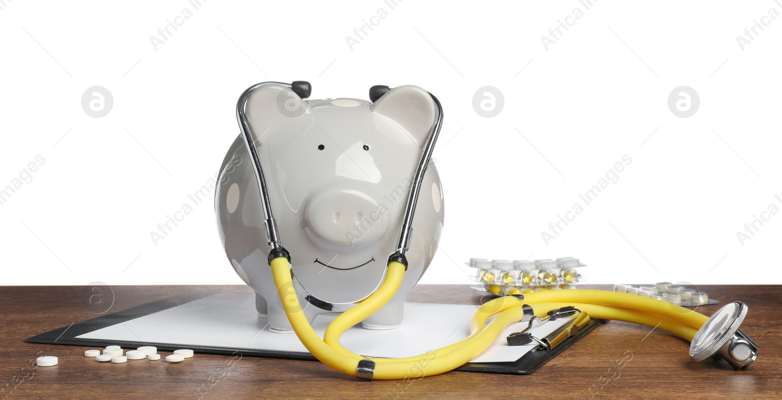 Photo of Piggy bank with stethoscope, clipboard and pills on wooden table against white background. Medical insurance