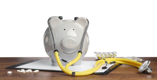 Photo of Piggy bank with stethoscope, clipboard and pills on wooden table against white background. Medical insurance