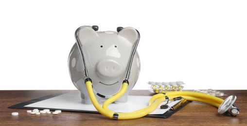 Piggy bank with stethoscope, clipboard and pills on wooden table against white background. Medical insurance