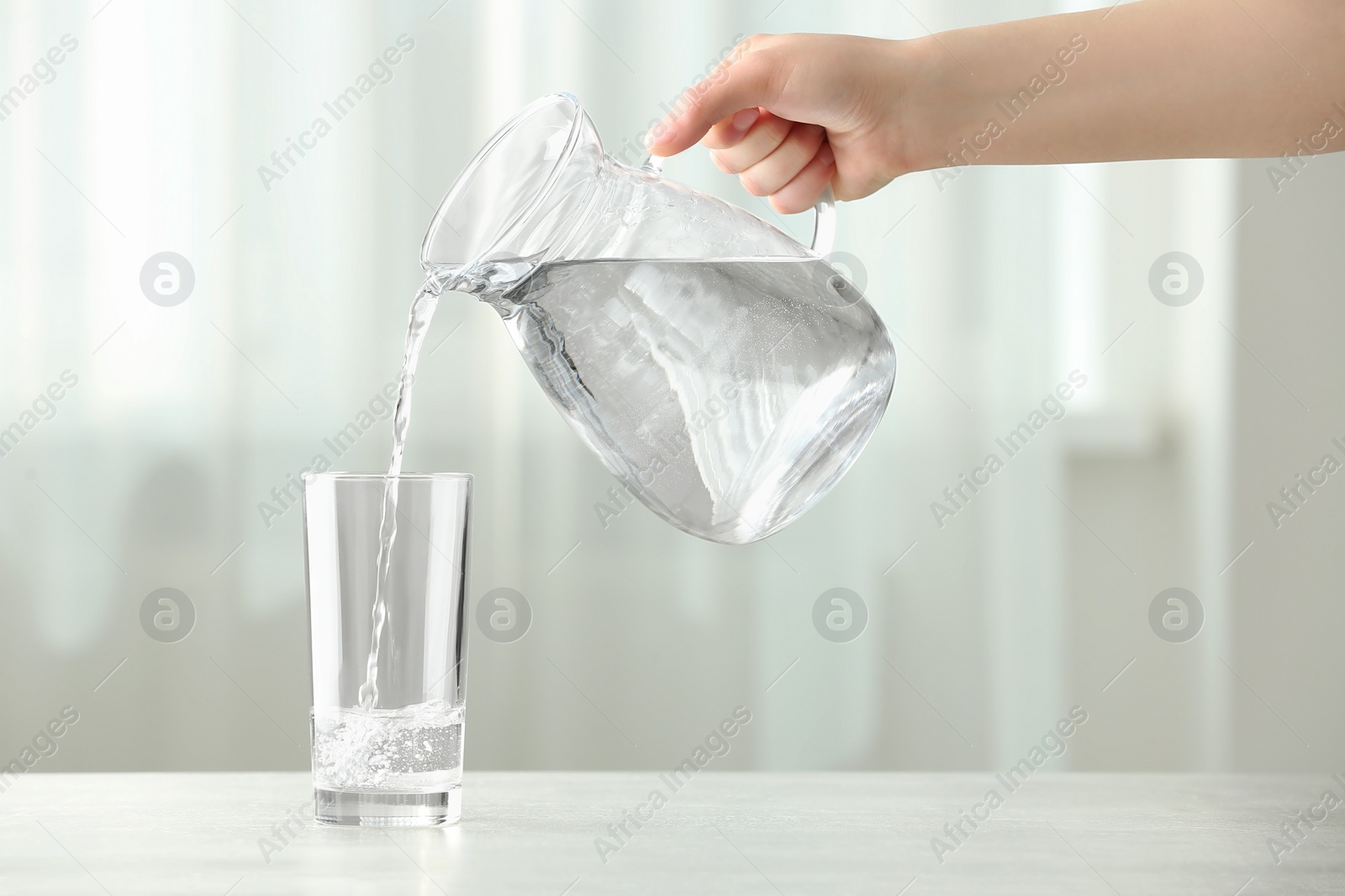 Photo of Woman pouring water from jug into glass at white table indoors, closeup