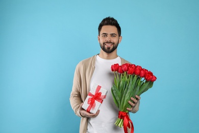 Photo of Happy man with red tulip bouquet and gift box on light blue background. 8th of March celebration