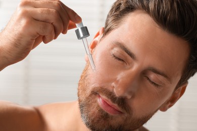 Handsome man applying cosmetic serum onto his face indoors, closeup
