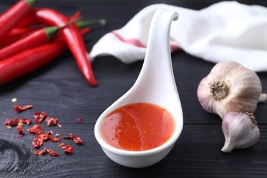 Photo of Spicy chili sauce in spoon, peppers and garlic on black wooden table, closeup