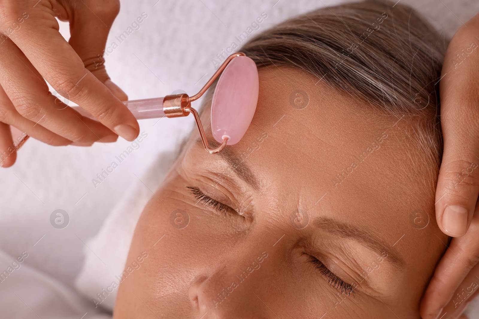 Photo of Woman receiving facial massage with rose quartz roller in beauty salon, closeup