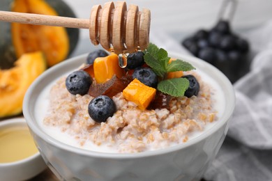 Photo of Adding honey to tasty wheat porridge with pumpkin, dates and blueberries in bowl, closeup
