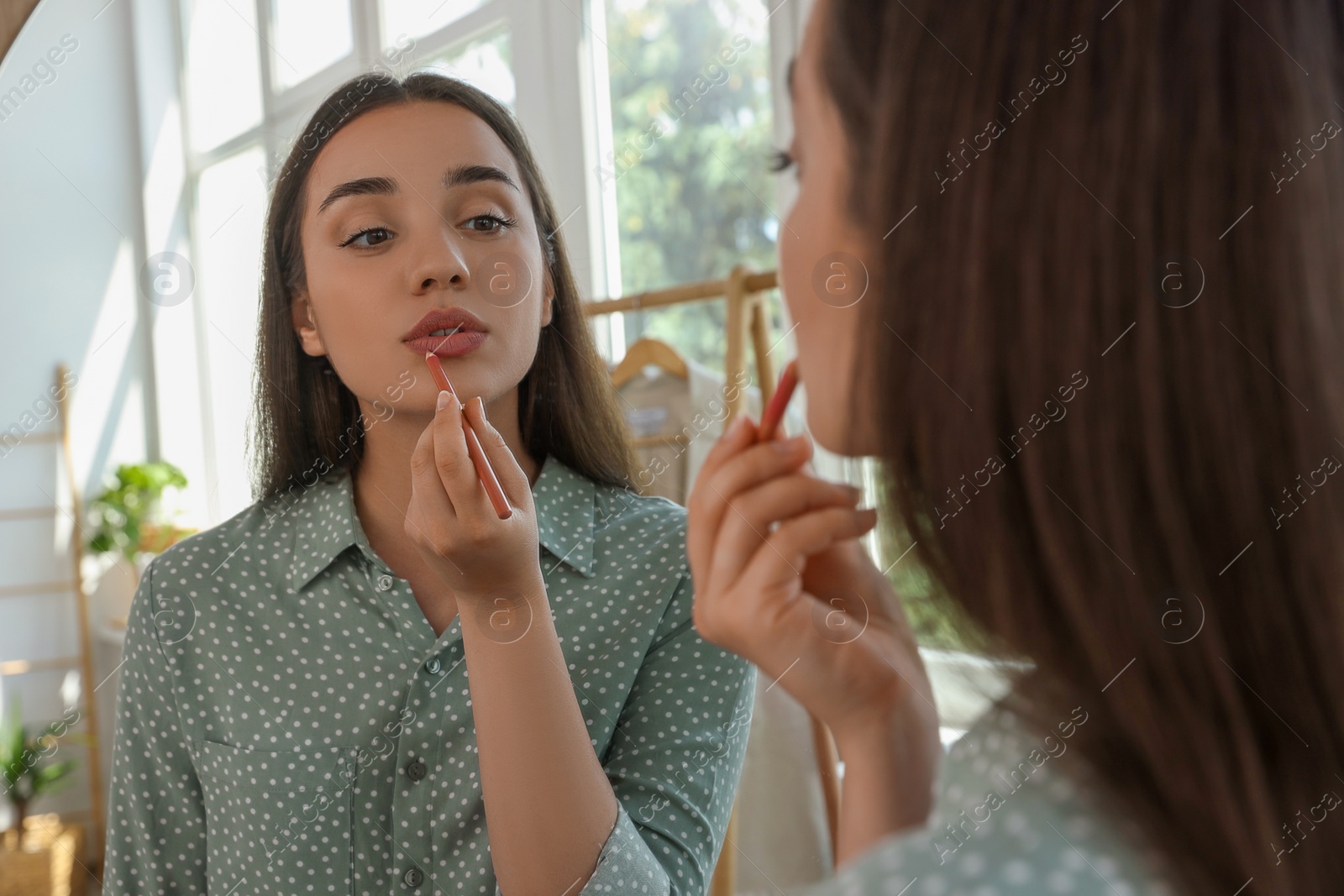 Photo of Beautiful young woman applying cosmetic pencil on lips near mirror indoors