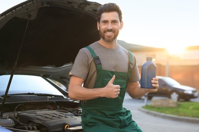 Photo of Smiling worker holding blue container of motor oil and showing thumbs up near car outdoors