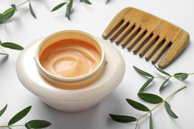 Photo of Jar of hair care cosmetic product, wooden comb and green leaves on white background, closeup