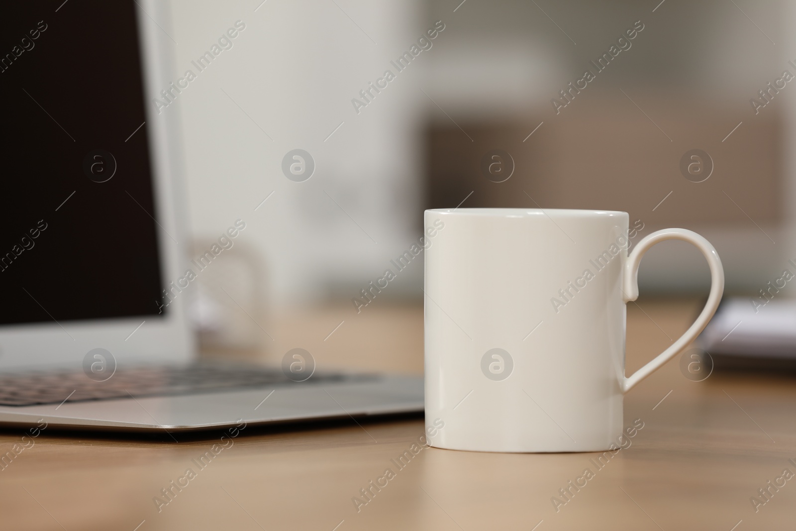 Photo of White ceramic mug and laptop on wooden table at workplace. Space for text